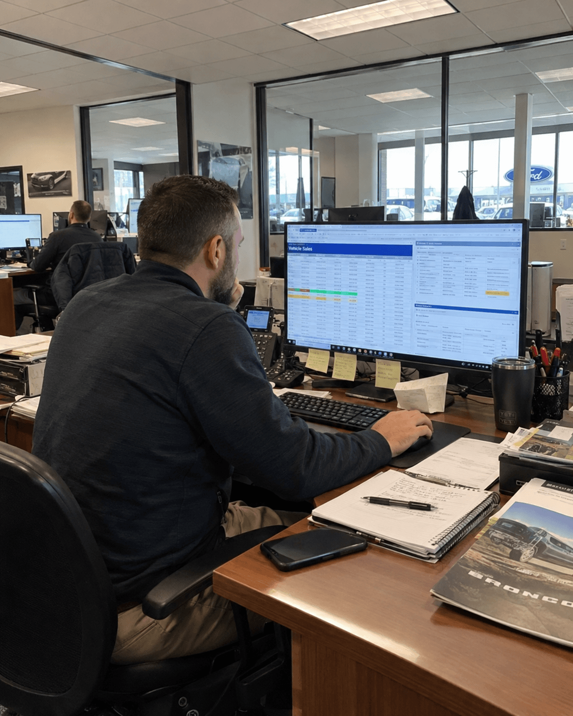 A business employee at his desk reviewing a vehicle sales table on a large monitor